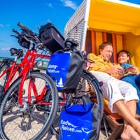 Two cyclists relaxing in a yellow-orange beach chair on the Baltic Sea beach in Wustrow, with red bicycles parked beside them under blue sky.