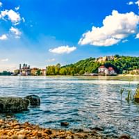 View across the Danube River in Passau with pebble shore, historic buildings and cathedral on the left, and a hilltop fortress under blue sky.