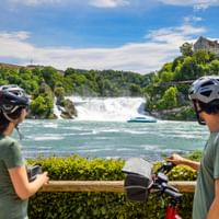 Two cyclists with helmets viewing Rhine Falls from a viewing platform. A tour boat crosses the turquoise water below the wide waterfall.
