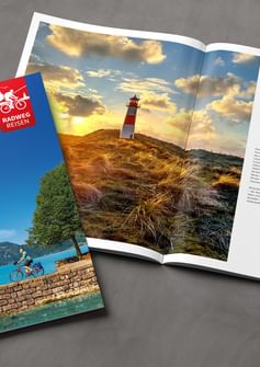 Radweg Reisen Katalog 2026 mit Cover von Radfahrern am See und aufgeschlagener Seite mit Nordsee-Leuchtturm bei Sonnenuntergang.