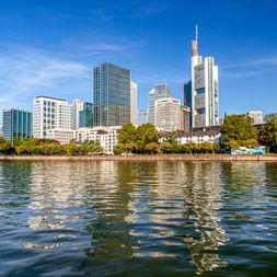 Frankfurter Skyline mit modernen Wolkenkratzern und Bürotürmen, die sich im Main spiegeln. Grüne Bäume säumen das Flussufer unter blauem Himmel.