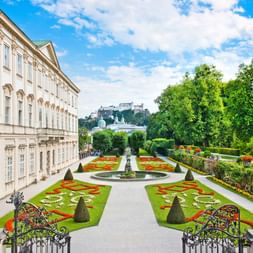 Schloss Mirabell mit barockem Garten mit bunten Blumenbeeten, Brunnen und geschnittenen Hecken. Festung Hohensalzburg auf dem Hügel sichtbar.