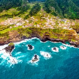 Luftaufnahme des Dorfes Seixal auf einer felsigen Halbinsel auf Madeira, umgeben von türkisfarbenem Meer und grünen Berghängen.