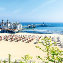 Sandiger Ostseestrand mit Reihen bunter Strandkörbe und einer historischen Seebrücke, die sich ins blaue Meer erstreckt.