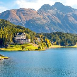 Waldhaus am See Hotel auf einer Halbinsel im St. Moritz See, umgeben von Wäldern und Alpenbergen unter blauem Himmel.