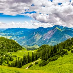 Malerische Berglandschaft bei Kochel am See mit grünen Almwiesen, Nadelwäldern und Herzogstand-Gebirgskette unter dramatischen Wolken.