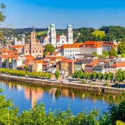 Panoramablick auf Passau entlang der Donau mit historischen Gebäuden, Kirchtürmen und orangefarbenen Dächern vor grünen Hügeln.