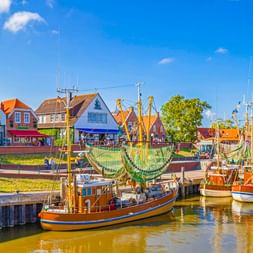 Traditionelle Fischerboote mit Netzen im Hafen von Greetsiel. Bunte Häuser mit roten Dächern säumen das Ufer unter blauem Himmel.