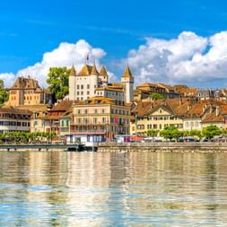 Blick auf das Schloss Nyon mit weißen Türmen am Genfersee. Bunte historische Gebäude säumen die Uferpromenade unter blauem Himmel.