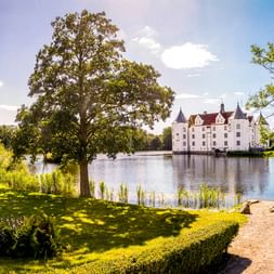 Weißes Schloss Glücksburg mit roten Dächern auf einem See, umgeben von grünen Rasenflächen, Bäumen und einem Kiesweg unter blauem Himmel.