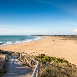 Holzsteg mit Seilgeländer führt durch Dünengras zu einem breiten Sandstrand an der Costa de la Luz. Blaues Meer mit Wellen und Küstenort sichtbar.