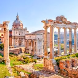 Antike römische Säulen und Ruinen am Forum Romanum mit barocker Kirchenkuppel im Hintergrund. Grüne Rasenflächen und archäologische Reste unter blauem Himmel.