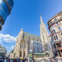 Stephansdom mit seinem hohen Turm und gemustertem Dach in der Wiener Innenstadt, umgeben von modernen und historischen Gebäuden unter blauem Himmel.