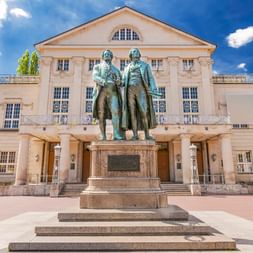 Deutsches Nationaltheater in Weimar mit Goethe-Schiller-Denkmal davor. Das klassizistische Gebäude hat einen dreieckigen Giebel und weiße Säulen.