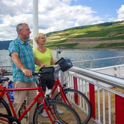 Paar mit rotem Tourenrad auf Rheinfähre, Blick auf terrassierte Weinberge am Hang. Blaue Fahrradtaschen am Rad.