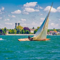Holzsegelboot mit weißem Segel auf dem Bodensee. Friedrichshafener Uferpromenade mit Kirchtürmen und Gebäuden im Hintergrund unter blauem Himmel.