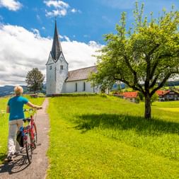 Radfahrer schiebt Fahrrad auf Weg durch grüne Wiese zur weißen Kirche mit spitzem Turm im Allgäu, mit Bergen im Hintergrund.