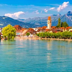 Blick auf Interlaken mit türkisfarbener Aare im Vordergrund, historischer Altstadt mit Kirchturm und Schweizer Alpengipfeln unter blauem Himmel.