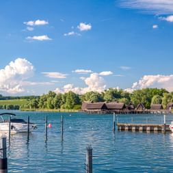 Hafen am Bodensee mit Booten und Holzsteg. Historische Pfahlbauten sind über dem Wasser unter blauem Himmel mit weißen Wolken sichtbar.