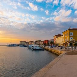 Uferpromenade in Poreč, Kroatien mit bunten Gebäuden, vertäuten Booten und Spaziergängern bei Sonnenuntergang unter dramatischem Wolkenhimmel.