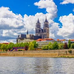 Der Magdeburger Dom mit zwei Türmen erhebt sich über der Elbe. Steinmauer, Bäume und Wohngebäude unter blauem Himmel mit Wolken sichtbar.