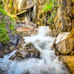 Rushing white water flows through the narrow Lötzklamm gorge in Austria. Moss-covered rock walls frame the stream, with wooden walkways visible above.