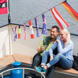 Paar sitzt auf Holzbank auf dem Deck des Schiffs Magnifique II mit internationaler Flaggenkette und Wasser im Hintergrund.