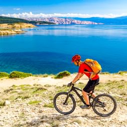Cyclist in red jersey with orange backpack riding mountain bike on coastal path in Croatia with blue sea and mountains in background.