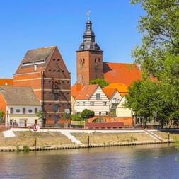 Blick auf die Havelberger Altstadtinsel über die Havel mit historischen Gebäuden mit orangefarbenen Ziegeldächern und einem markanten Backsteinkirchturm.