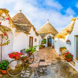 Weiß getünchte Trulli-Häuser mit konischen Steindächern in Alberobello. Ein Fahrrad mit roten Blumen steht in einer engen Gasse mit Topfpflanzen.