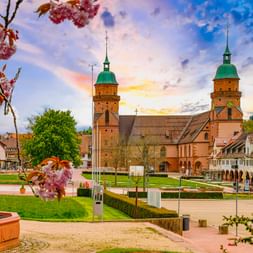 Freudenstädter Stadtkirche mit zwei grünen Kuppeltürmen am Marktplatz. Rosa Kirschblüten rahmen die Ansicht unter blauem Himmel.