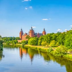 Blick auf Schloss Johannisburg mit roten Sandsteintürmen über den Main in Aschaffenburg, umgeben von grünen Bäumen unter blauem Himmel.