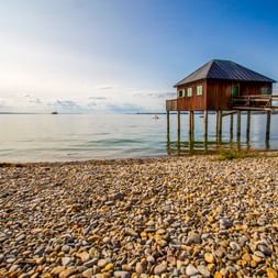 Hölzernes Badehaus auf Stelzen über dem Bodensee in Bregenz. Kieselstrand im Vordergrund, ruhiges Wasser und blauer Himmel mit Wolken im Hintergrund.