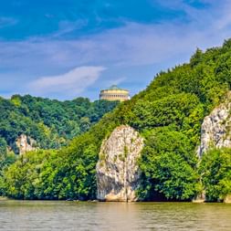 Befreiungshalle auf bewaldetem Hügel über der Donau bei Kelheim. Kalksteinfelsen ragen aus dem Wasser inmitten üppiger Vegetation.