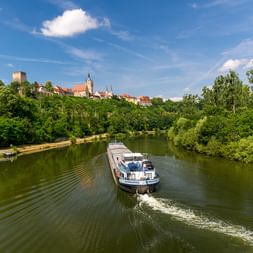 Frachtschiff auf dem Neckar mit Bad Wimpfens historischer Kaiserpfalz und Kirchturm auf dem Hügel über grünen Bäumen unter blauem Himmel.