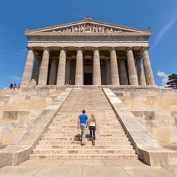 Zwei Personen steigen Steintreppen zur Walhalla an der bayerischen Donau hinauf. Der neoklassizistische Tempel zeigt dorische Säulen und Giebel.