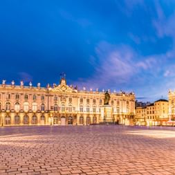 Place Stanislas in Nancy bei Abenddämmerung mit beleuchteten Barockgebäuden, verzierten Straßenlaternen und Kopfsteinpflaster unter blauem Himmel.
