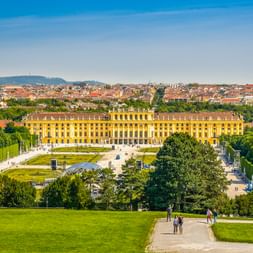 Gelbes Schloss Schönbrunn in Wien, umgeben von formalen Gärten und Bäumen, mit Besuchern auf Wegen und Wiener Stadtbild im Hintergrund.