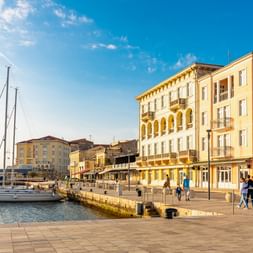 Hafenpromenade von Poreč mit Segelbooten, Fußgängern und bunten mediterranen Gebäuden mit Arkadenfassaden unter blauem Himmel.