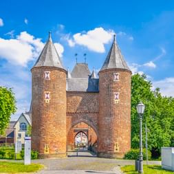 Klever Tor, ein mittelalterliches Backsteintor mit zwei runden Türmen und Spitzdächern in Xanten, umgeben von grünen Bäumen unter blauem Himmel.