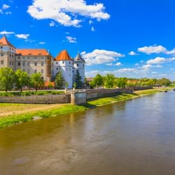 Schloss Hartenfels in Torgau mit orangen Dächern und rundem Turm an der Elbe. Steinbrücke verbindet das Schloss mit grünen Ufern unter blauem Himmel.