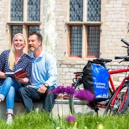 Zwei Radfahrer mit einem roten Tourenrad sitzen auf einer Bank in Middelburg, mit historischem Gebäude und lila Blumen im Hintergrund.