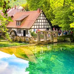 Kristallklarer türkisfarbener Blautopf in Blaubeuren mit Fachwerkhaus und Holzbrücke. Besucher auf der Brücke, umgeben von grünen Bäumen.