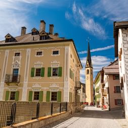 Colorful historic buildings with green shutters line a cobblestone street leading to a church with a tall spire in an Austrian old town under blue sky.