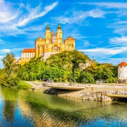 Das gelbe barocke Stift Melk mit Zwillingstürmen thront auf einem grünen Hügel an der Donau. Eine geschwungene Promenade und Wohnhäuser säumen das Ufer.