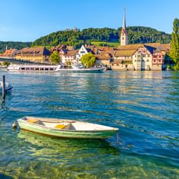 Blick auf Stein am Rhein am Rheinufer mit Booten im Vordergrund, historischen Gebäuden, Kirchturm und bewaldeten Hügeln im Hintergrund.