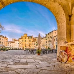 Blick durch Steinbogen auf die Piazza dell'Anfiteatro in Lucca. Rotes Fahrrad lehnt an Bogenpfeiler, ovaler Platz mit historischen Gebäuden.