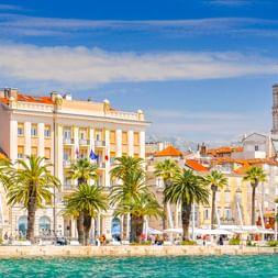 Split waterfront promenade with palm trees, historic buildings with orange roofs, and the cathedral bell tower under a blue sky.