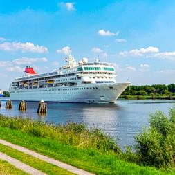 Weißes Kreuzfahrtschiff auf dem Nord-Ostsee-Kanal in Schleswig-Holstein. Grüner Radweg entlang des Wasserwegs mit Holzpfählen im Wasser.