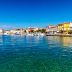 Poreč harbor with colorful historic buildings along the waterfront, boats moored in clear turquoise water under a blue sky.